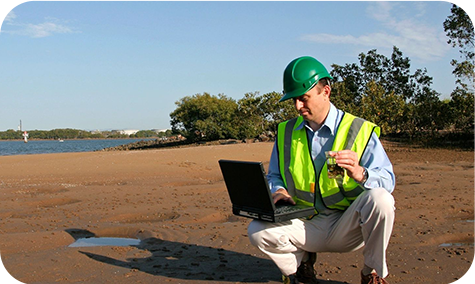 A man in yellow vest and hard hat using laptop on beach.